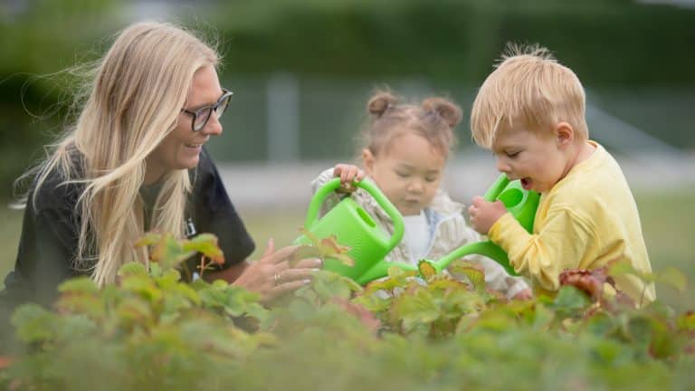 Dzieci i opiekunka sadzą rośliny na zewnątrz, korzystając z zielonych konewek.