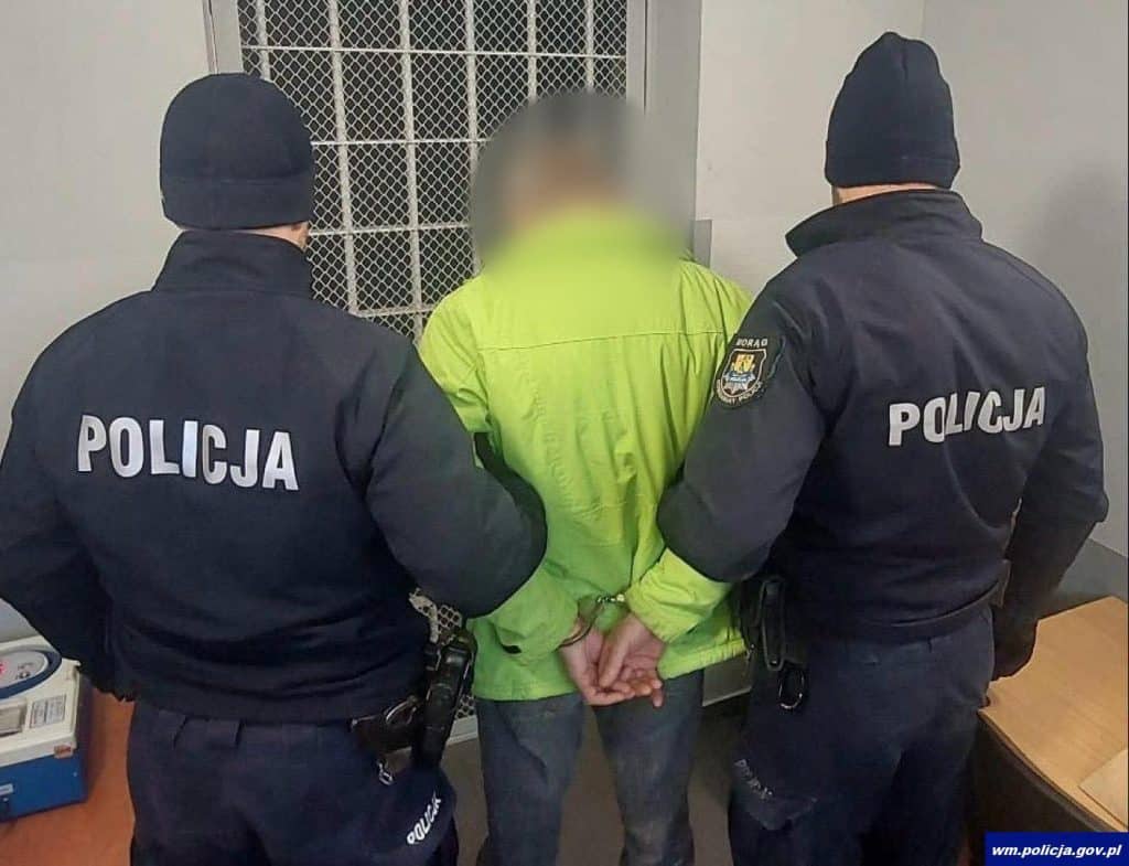 Policjanci zatrzymują mężczyznę w mieszkaniu, z tyłu widoczna klatka schodowa.