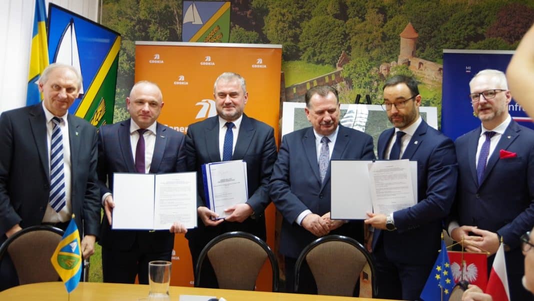 Grupa mężczyzn w formalnych ubraniach podczas podpisania umowy, za nimi banery i flagi, w tle krajobraz.