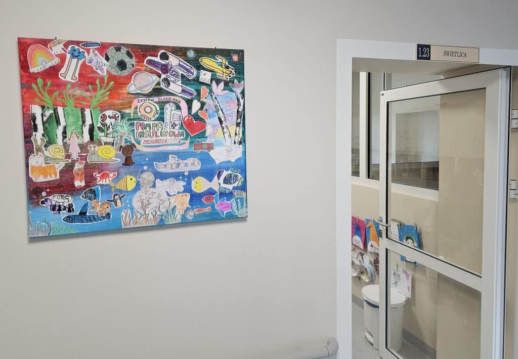 Kolorowa dziecięca sala szpitalna z rysunkami na ścianie i drzwiami do oddziału.