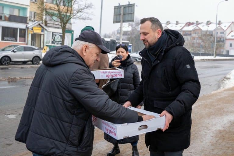 Prezydent Olsztyna rozdaje pączki na ulicy w zimowej scenerii, rozmawia z mieszkańcami.