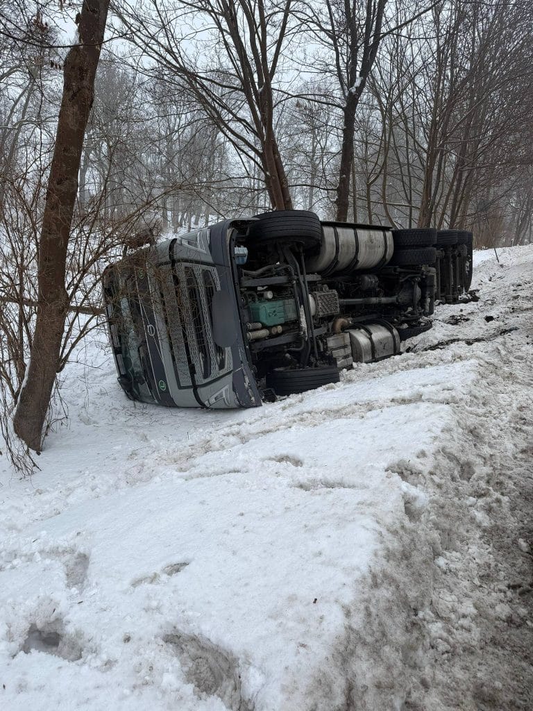 Rozbity ciężar&oacute;wka leży na boku na śnieżnej drodze w lesie.