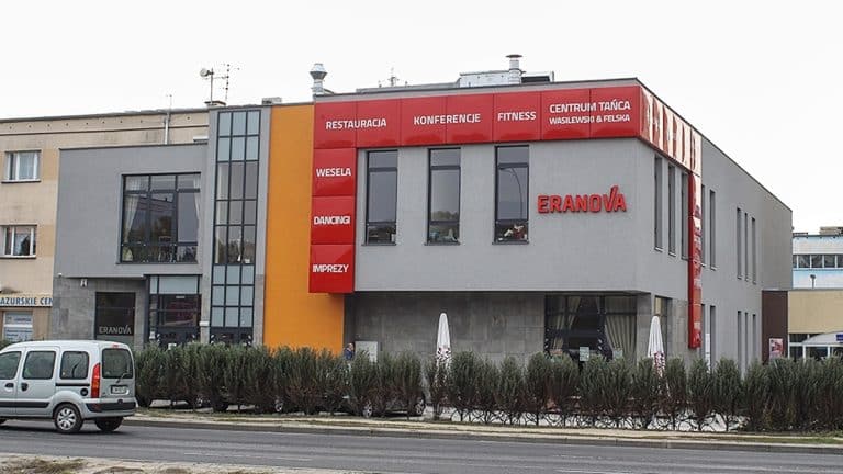 Nowoczesny budynek centrum eventowego w centrum Olsztyna z dużymi oknami i czerwonymi tablicami informacyjnymi.