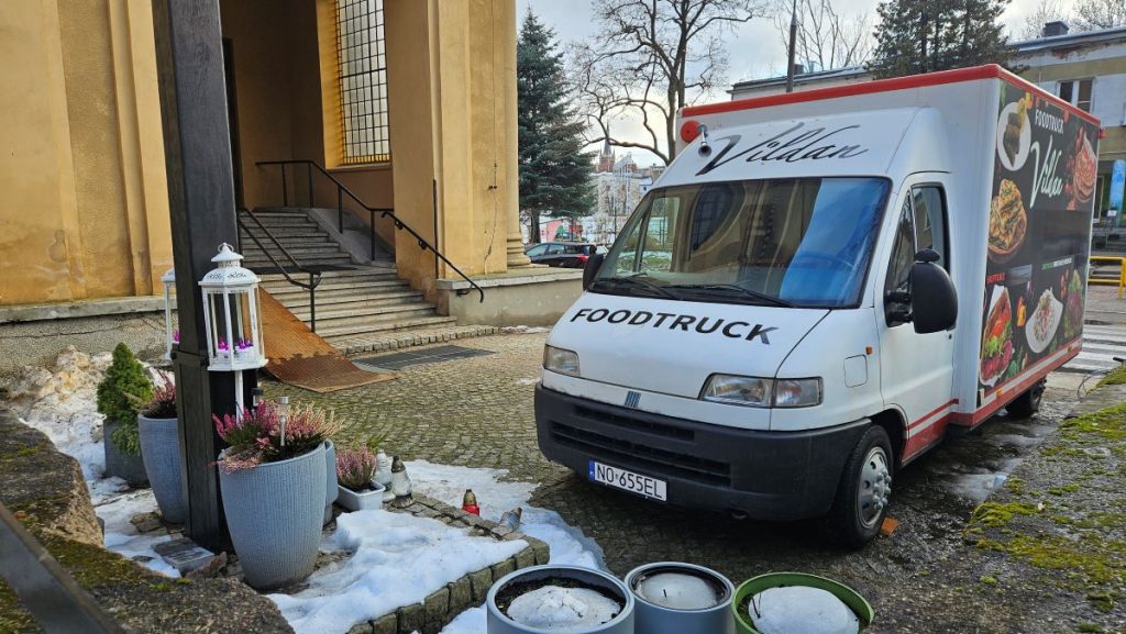 Food truck z kawą i jedzeniem na tle parafii na Zatorzu, w pobliżu kwiatów i śniegu.