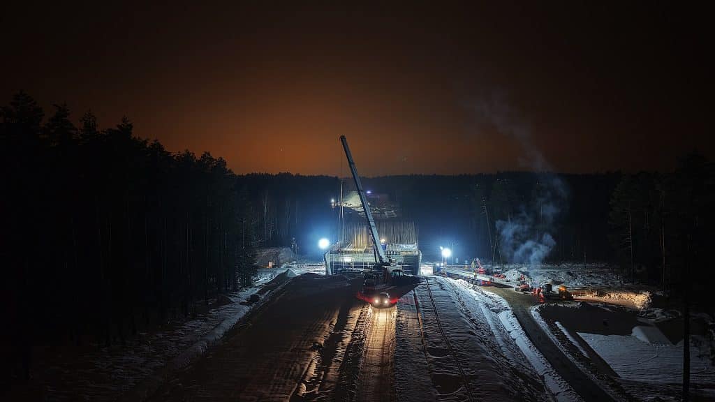 Prace budowlane na drodze w nocy, z widocznym ciężkim sprzętem i oświetleniem na placu budowy.