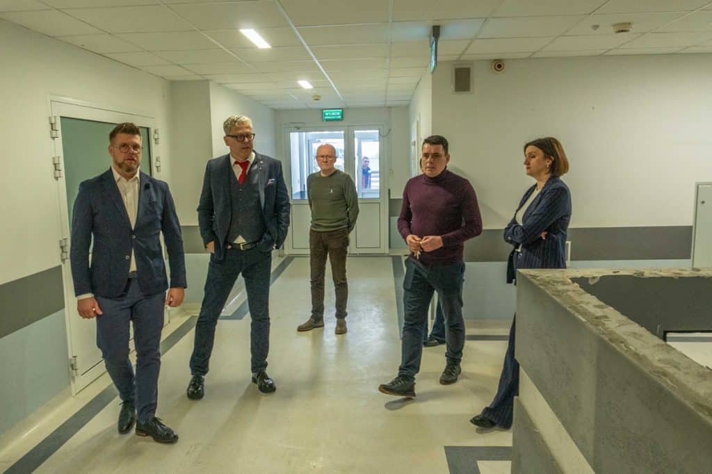 Grupa os&oacute;b w korytarzu szpitala, w tle drzwi wejściowe, na podłodze linie i oznaczenia.