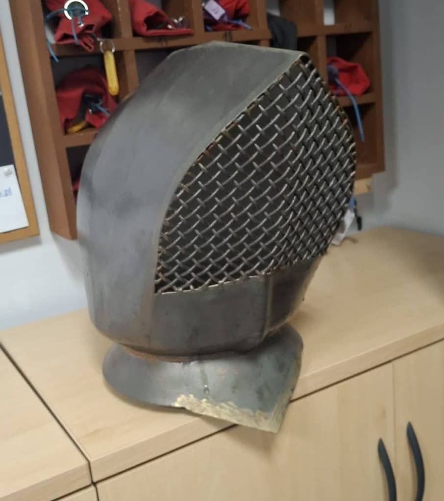 Metalowa głowa rycerza z zabytkowej figury, odłączona od ciała, ustawiona na stole.