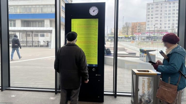 Dwie osoby czekają przy automacie biletowym na dworcu, z ekranem wyświetlającym komunikaty o awarii systemu.