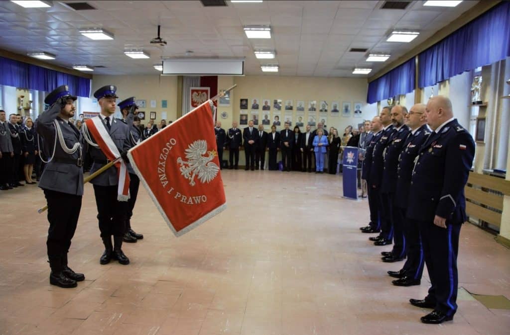 Policjanci podczas uroczystości, w tle portrety i flagi, w centrum tr&oacute;jka funkcjonariuszy z flagą.