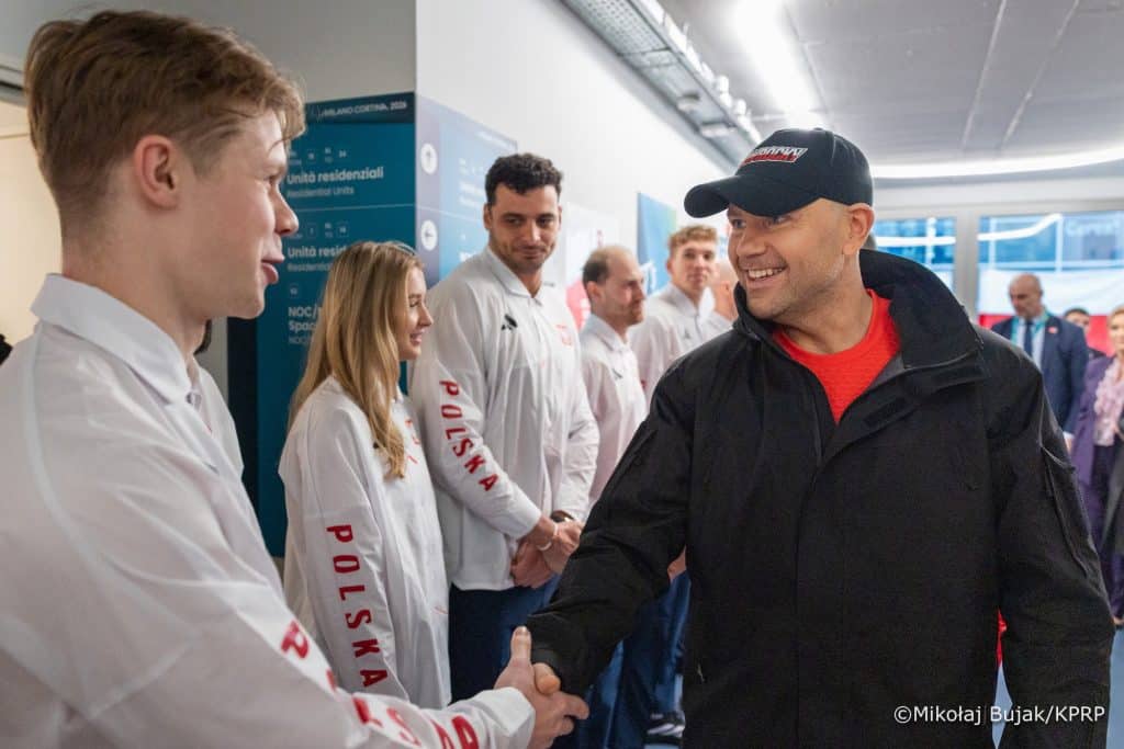 Mężczyzna uśmiecha się i podaje rękę innemu mężczyźnie w tle, w tle grupa ludzi w białych koszulach.