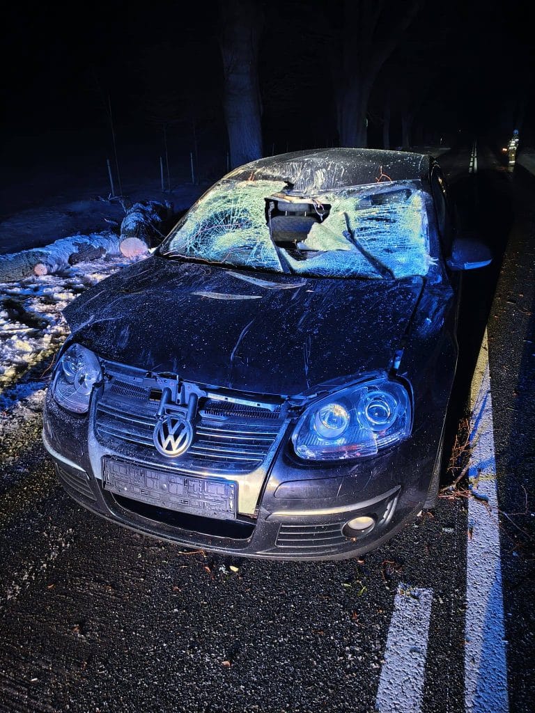 Uszkodzony samoch&oacute;d z rozbitym przednim i uszkodzonym dachem na drodze, w tle nocne oświetlenie i służby ratunkowe.
