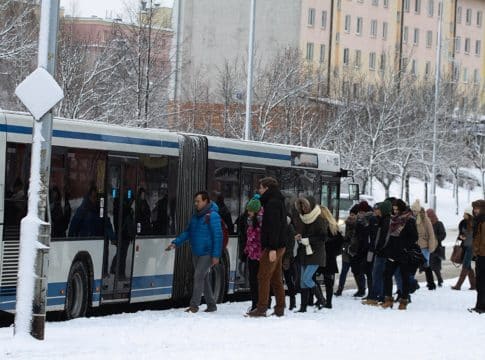 Ludzie wsiadają do autobusu na przystanku w zimowej scenerii, za nimi bloki mieszkalne i zamarznięte drzewa.