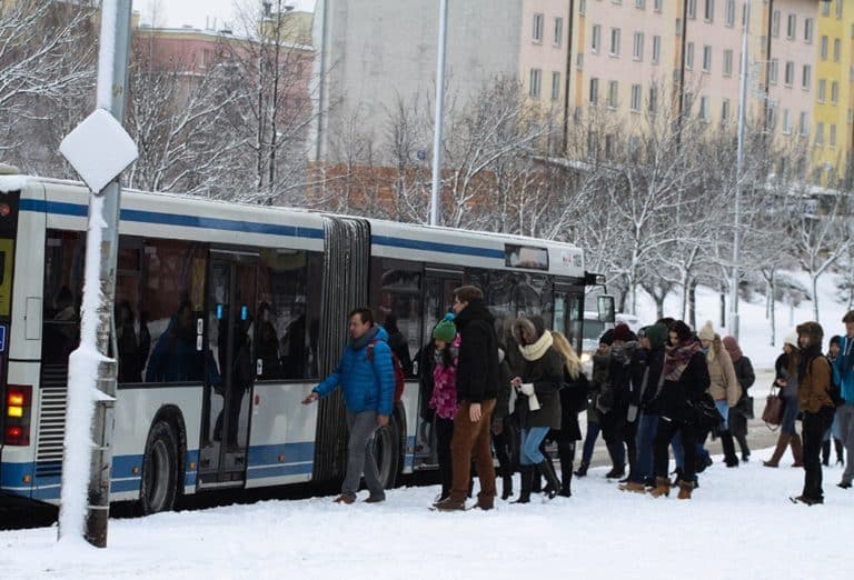 Ludzie wsiadają do autobusu na przystanku w zimowej scenerii, za nimi bloki mieszkalne i zamarznięte drzewa.