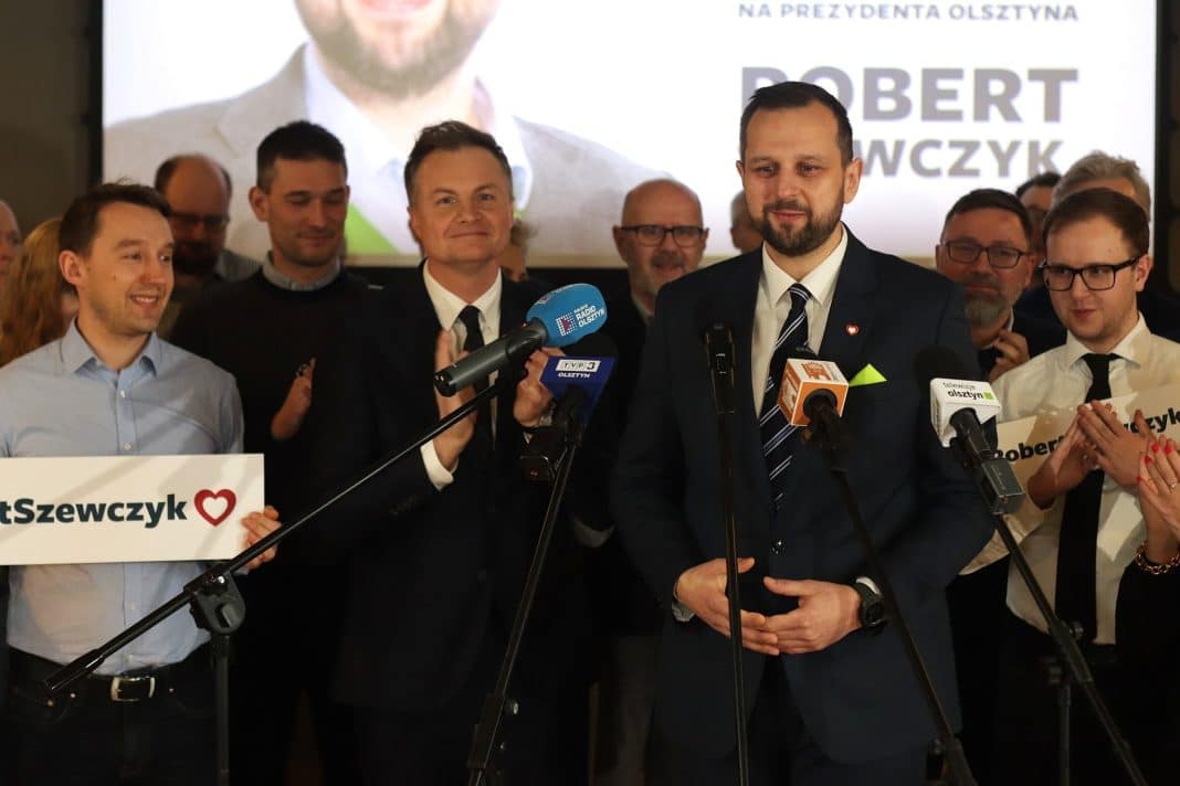Grupa ludzi na konferencji prasowej, w centrum mężczyzna w garniturze przy mikrofonach.