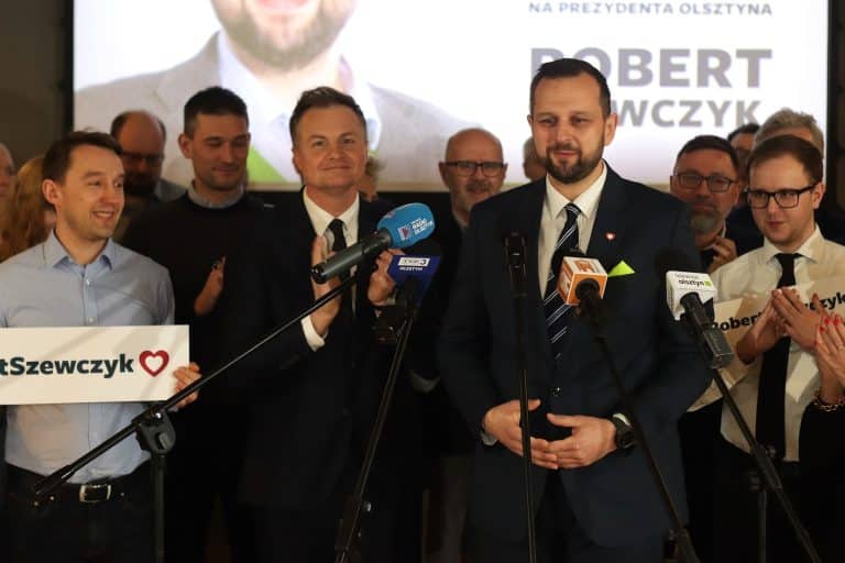 Grupa ludzi na konferencji prasowej, w centrum mężczyzna w garniturze przy mikrofonach.