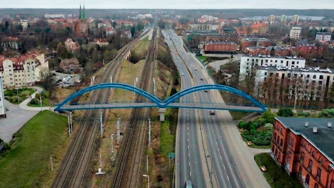 Widok na most nad torami kolejowymi i ulicami Olsztyna, z drzewami i budynkami w tle.