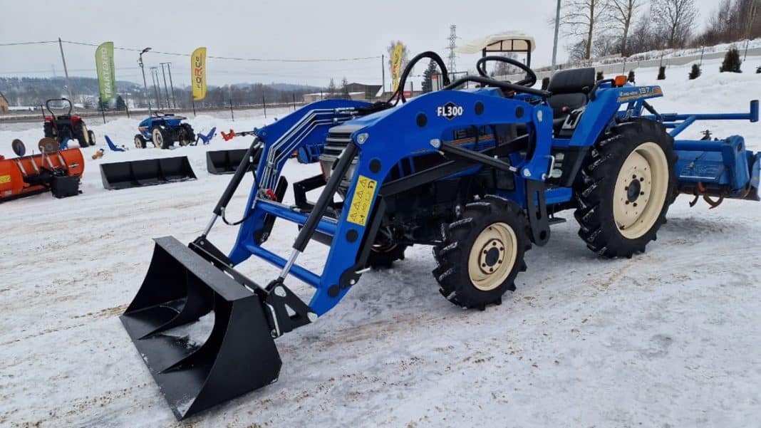 Niewielki, niebieski ciągnik japoński na śnieżnym terenie z innymi maszynami w tle.