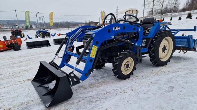 Niewielki, niebieski ciągnik japoński na śnieżnym terenie z innymi maszynami w tle.