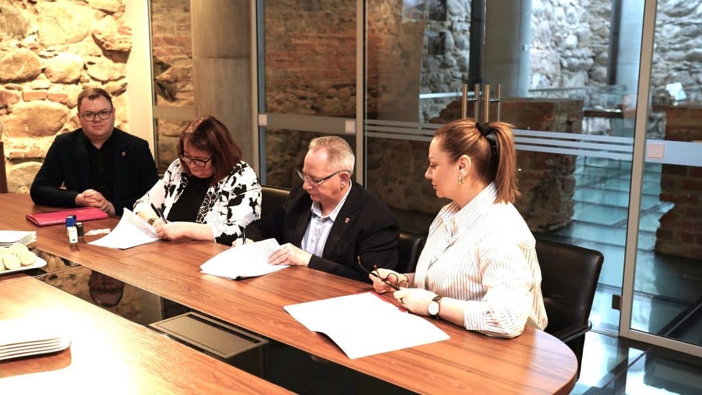 Zdjęcie przedstawia grupę czterech os&oacute;b siedzących przy stole podczas spotkania, z widokiem na kamienne ściany i szklane drzwi.