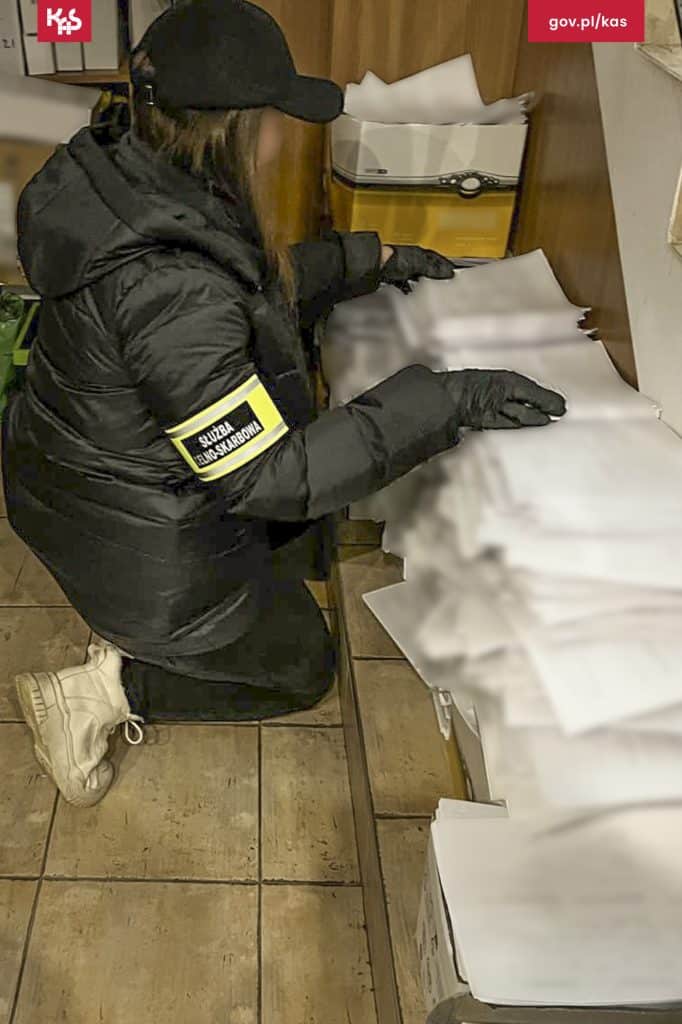 Funkcjonariuszka przeszukuje dokumenty na biurku, ubrana w czarną kurtkę i rękawiczki, w pomieszczeniu biurowym.