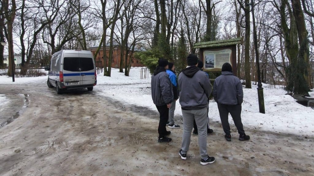 Grupa osadzonych ogląda tablicę informacyjną w parku zimą, w tle drzewa i samoch&oacute;d.