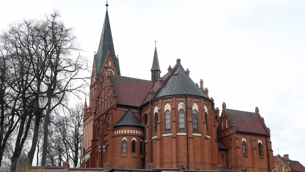 Zabytkowa bazylika w Gietrzwałdzie z ceglaną architekturą i wysoką wieżą, otoczona drzewami.