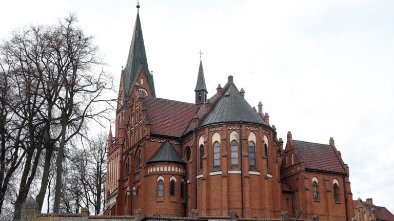 Zabytkowa bazylika w Gietrzwałdzie z ceglaną architekturą i wysoką wieżą, otoczona drzewami.
