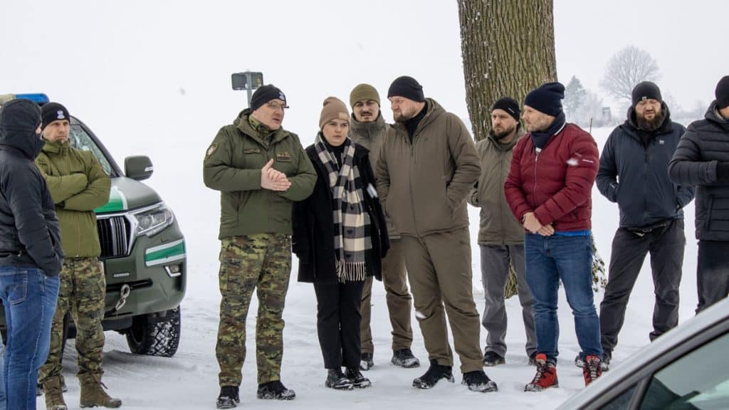 Grupa funkcjonariuszy i przedstawicieli służb na śnieżnym terenie, rozmawiających przy drzewie.
