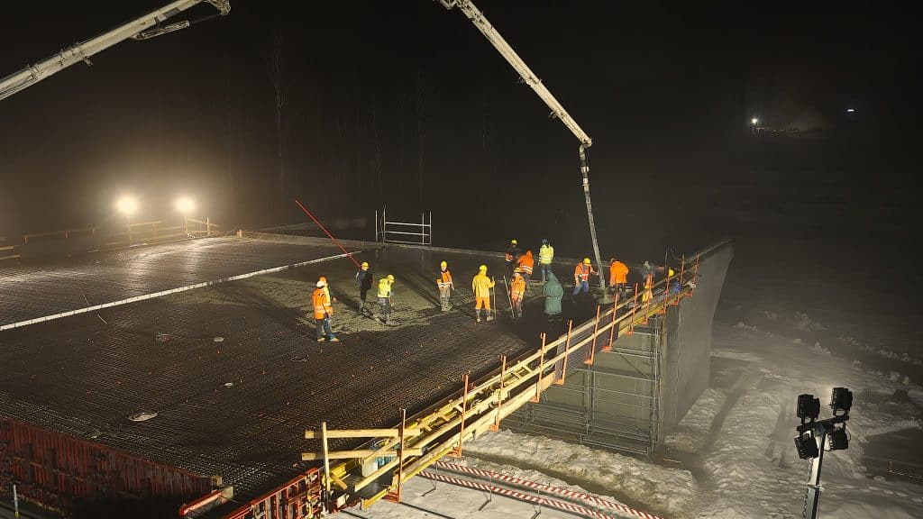 Prace budowlane na moście w nocy, z ciężkim sprzętem i robotnikami, w zimowych warunkach.