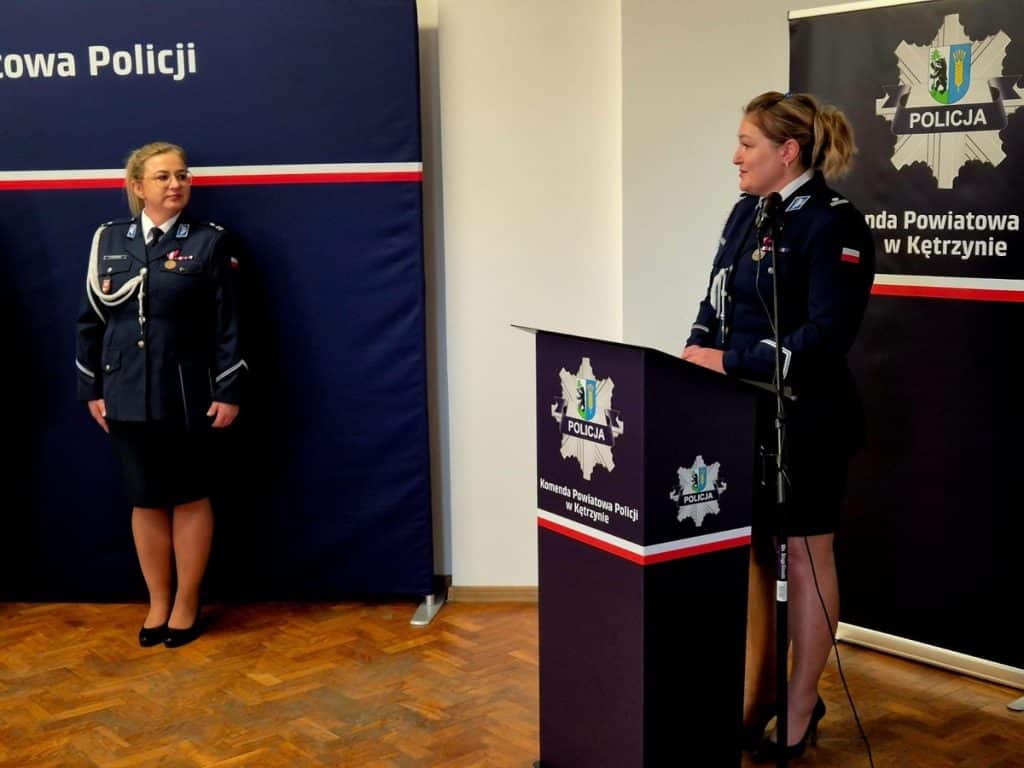 Dwie policjantki w uniformach podczas konferencji w pomieszczeniu z logo policji na tle.