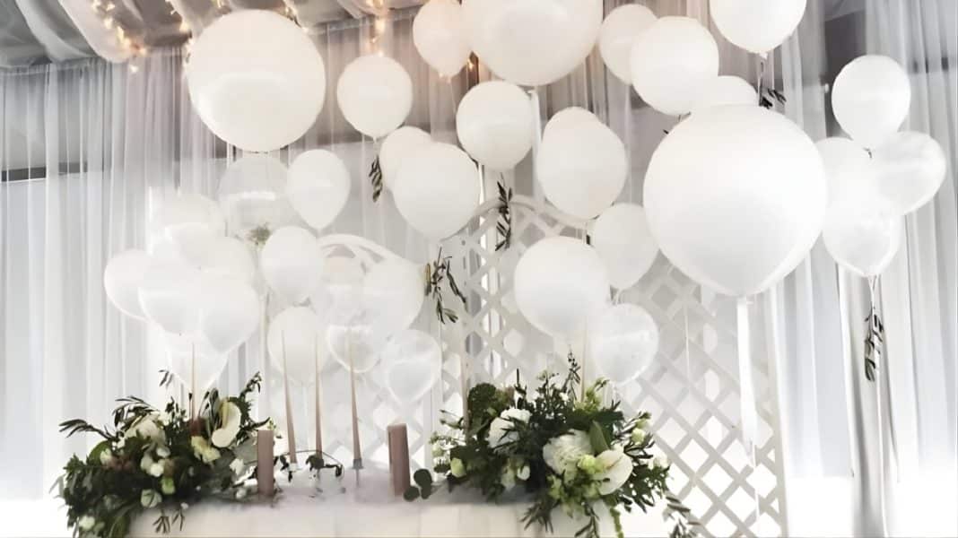 Dekoracja świąteczna z białymi balonami i kwiatami na tle białych zasłon.