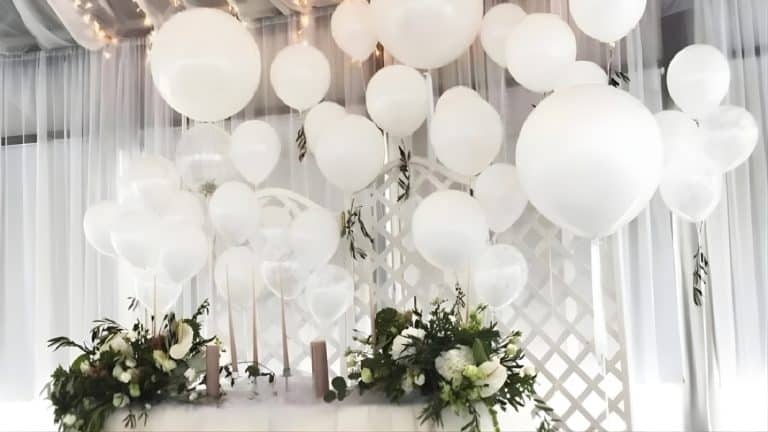 Dekoracja świąteczna z białymi balonami i kwiatami na tle białych zasłon.