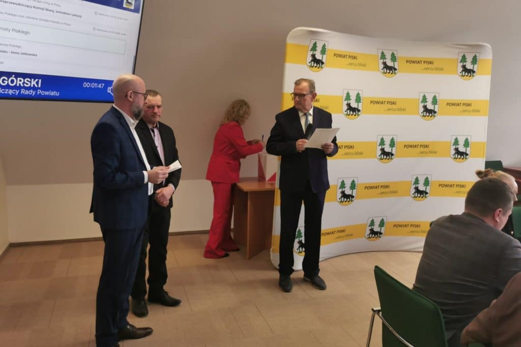 Uroczystość w powiecie piskim, nowy starosta odczytuje przem&oacute;wienie, w tle logo powiatu.