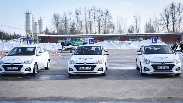 Trzy samochody szkoleniowe na placu egzaminacyjnym z tablicami L, w tle śnieg i drzewa.