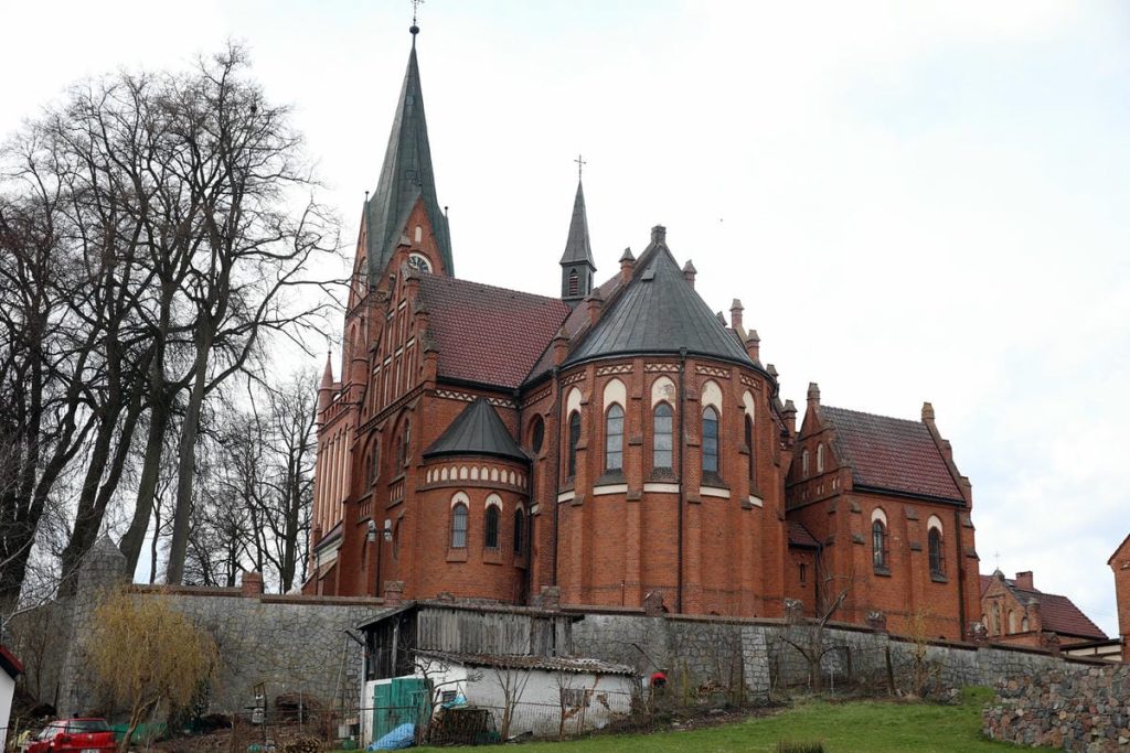 Zabytkowa bazylika w Gietrzwałdzie z widokiem na jej ceglane mury i wieże, otoczona drzewami i niskimi budynkami.
