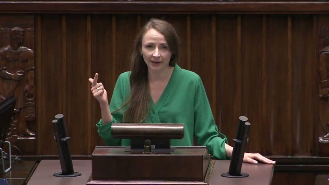 Kobieta przemawia podczas posiedzenia w drewnianej sali, gestykulując ręką.