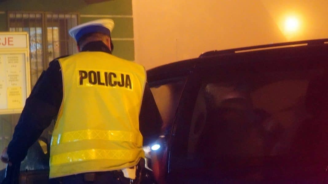 Policjant w kamizelce z napisem 'Policja' zatrzymuje samoch&oacute;d na parkingu w Ełku.