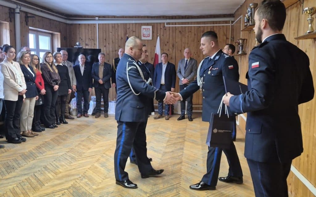 Policjanci podczas uroczystości, w tle grupa os&oacute;b i drewniane ściany.