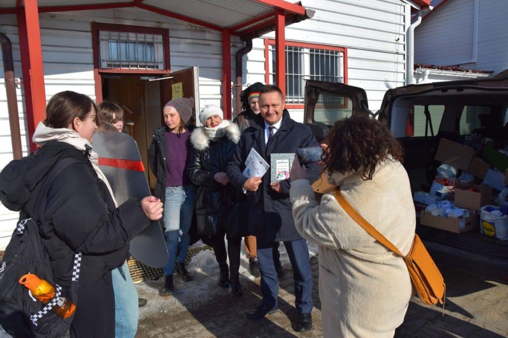 Grupa ludzi podczas akcji wymiany kubk&oacute;w na zewnątrz budynku, z samochodem pełnym kubk&oacute;w w tle.