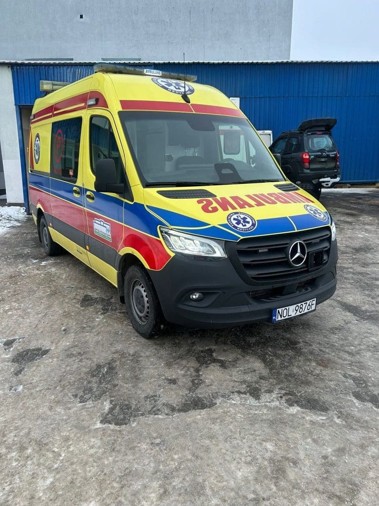 Ambulans pogotowia stojący na parkingu przed szpitalem, w tle budynek i inny samoch&oacute;d.