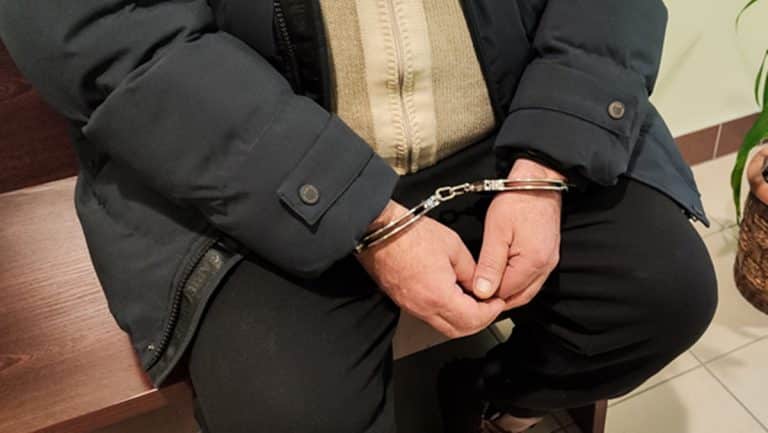 Zatrzymanie mężczyzny w rękawicach, z kajdankami na rękach, siedzącego na ławce.