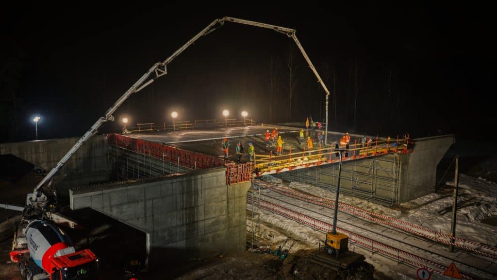 Prace budowlane na moście w nocy, z ciężkim sprzętem i robotnikami, oświetlonymi lampami.