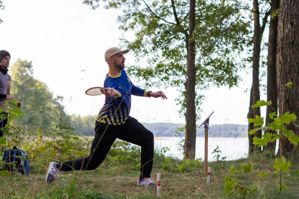 Mężczyzna rzuca disc golfem na tle drzew i jeziora podczas zimowych trening&oacute;w w Olsztynie.