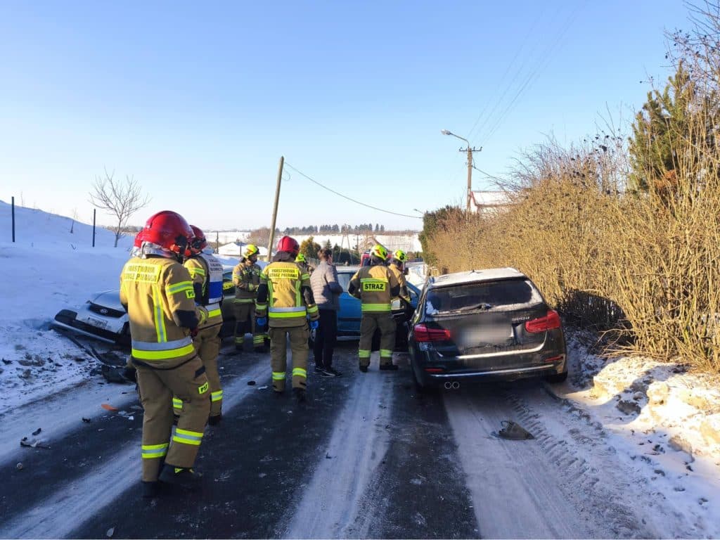 Zdjęcie przedstawia zderzenie dw&oacute;ch samochod&oacute;w na śliskiej, zaśnieżonej drodze, z obecnością strażak&oacute;w i policji.