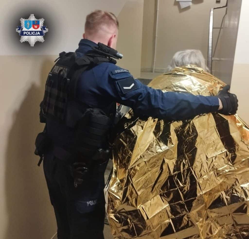 Policjant w kamizelce zabezpiecza osobę owiniętą w złotą folię w budynku.