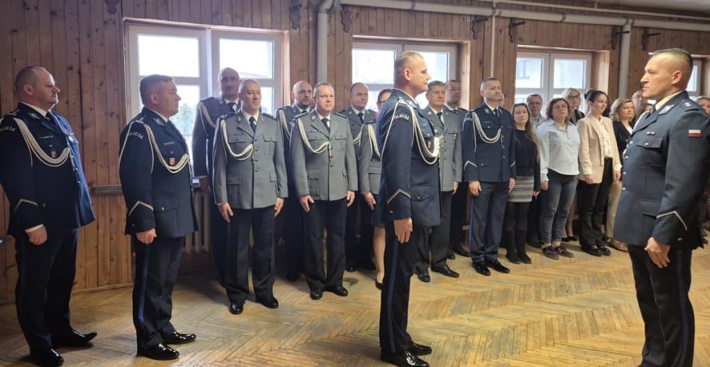 Policjanci podczas uroczystości w drewnianym pomieszczeniu, w tle ludzie w cywilnych ubraniach.
