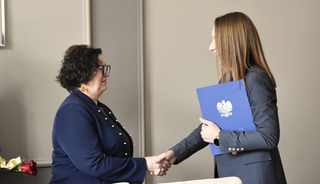 Dwie kobiety w urzędzie, jedna wręcza drugiej dokument w niebieskiej teczce z orłem na okładce.