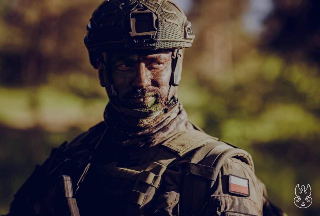 Żołnierz w kamuflażu i hełmie na tle natury, w słoneczny dzień.