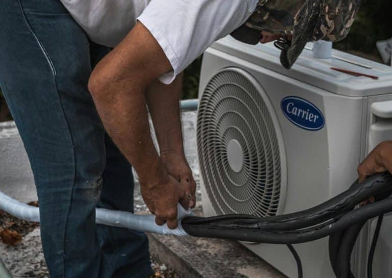 Pracownik serwisuje klimatyzator na zewnątrz budynku, podłączając przewody do urządzenia.