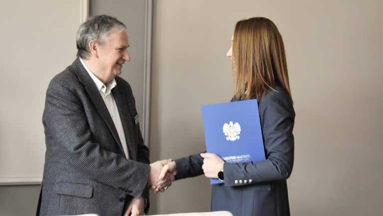 Urzędnik i kobieta podczas oficjalnego wręczenia dokumentu, w tle neutralna ściana.